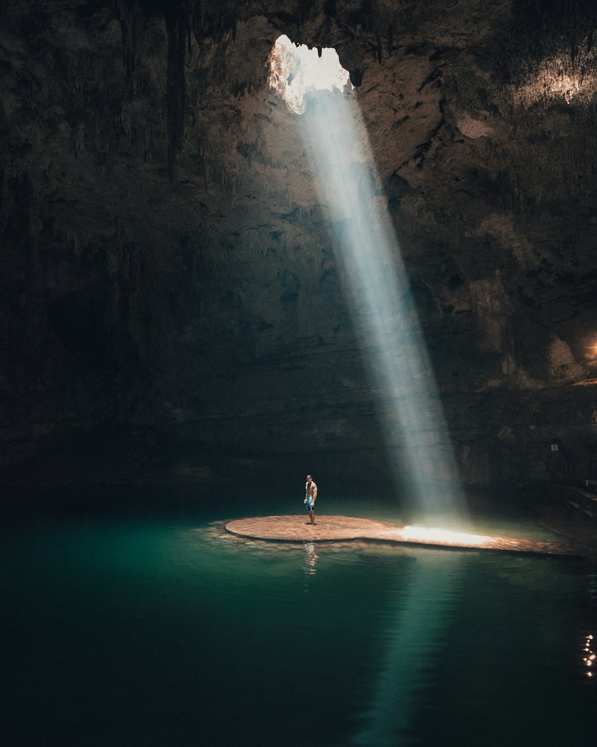 Person standing on a rock platform inside a dramatic cave cenote with a beam of light cutting through the water, Yucatan