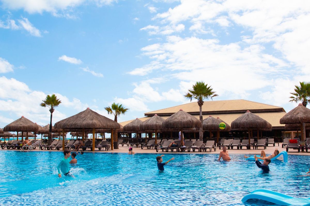 Families playing in a resort pool beside thatched palapas
